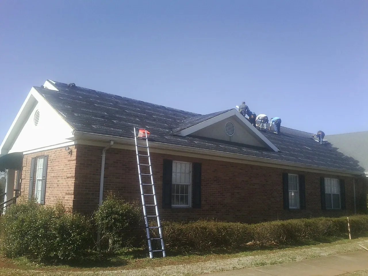 Crew tearing shingles off a brick ranch roof with a ladder set — Emergency Roof Repair work by our crew in Mount Ayr