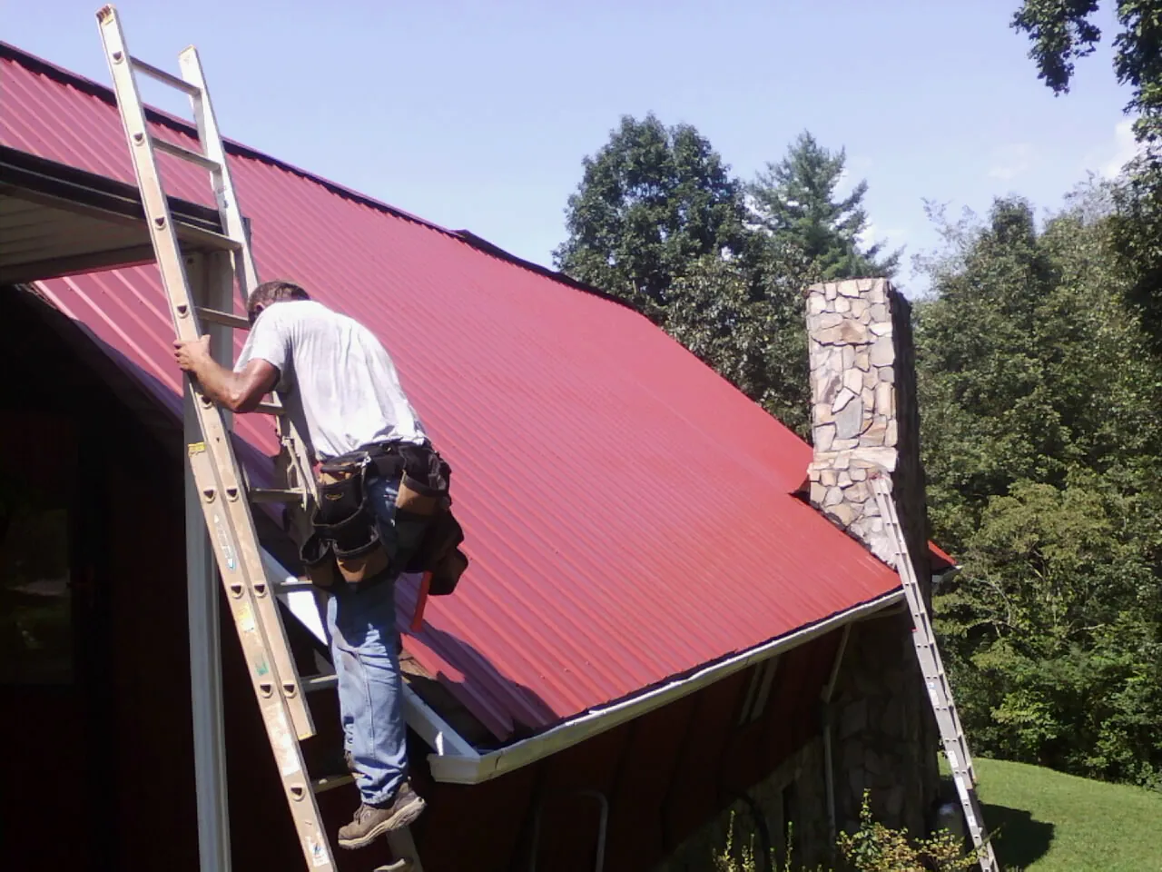 Roofer on a ladder beside a red metal roof and stone chimney — Soffit & Fascia Repair by Roofing Brothers in Mount Ayr